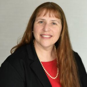 Smiling light skinned woman with auburn hair and wearing a black blazer, red shirt, and pearls