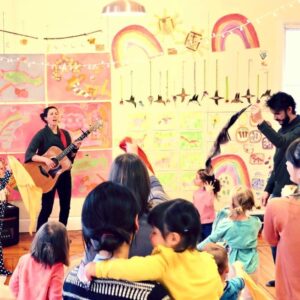 A woman in front of a wall full of childrens drawings playing guitar and singing while children watch and dance in front of her