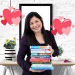 Light skinned, dark haired woman holding a stack of books with a table and floating drawn hearts behind her