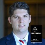 Light skinned dark haired young man wearing a blue suit and red and white striped tie next to the book The Power of Partisanship by Joshua Dyck